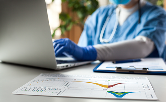 A medical professional wearing a stethoscope and latex gloves, typing into a laptop computer with printouts of charts